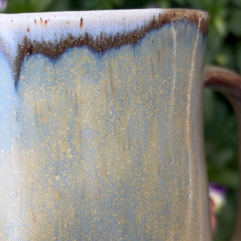 Handmade ceramic mug with brown drips and iridescent effects of gold and blue. Close up photo.