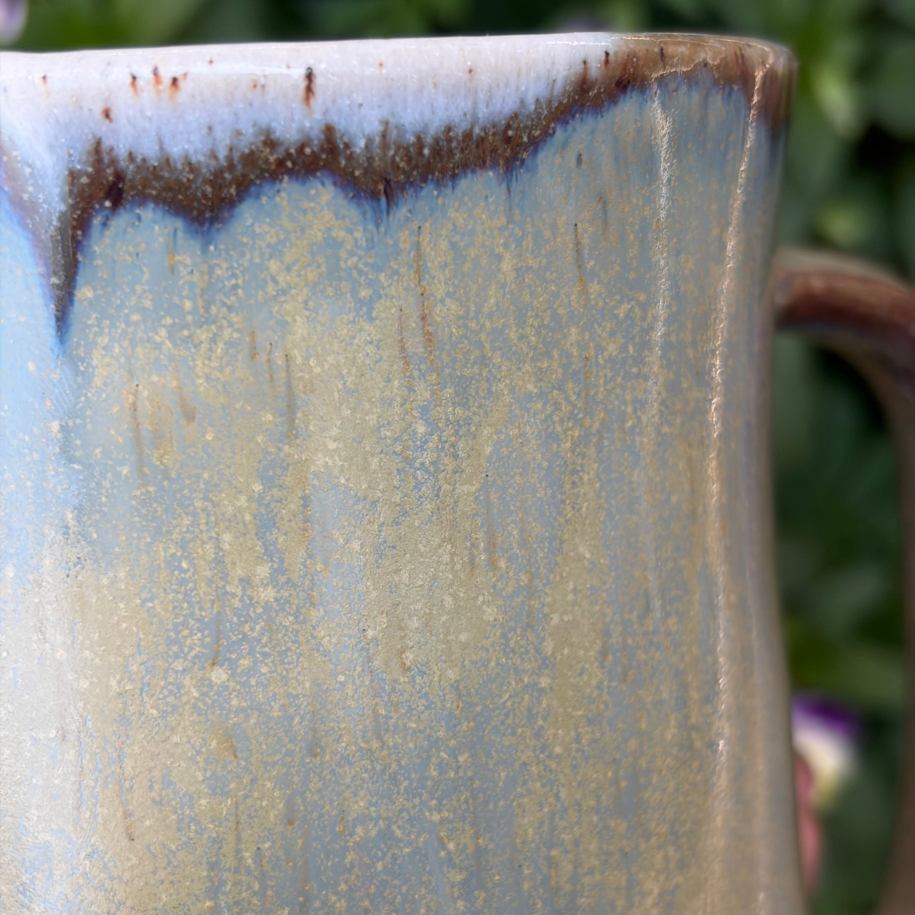 Handmade ceramic mug with brown drips and iridescent effects of gold and blue. Close up photo.